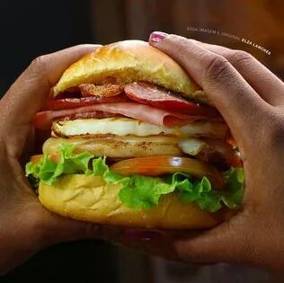 Mãos segurando um hambúrguer da Elza com bacon, ovo e alface